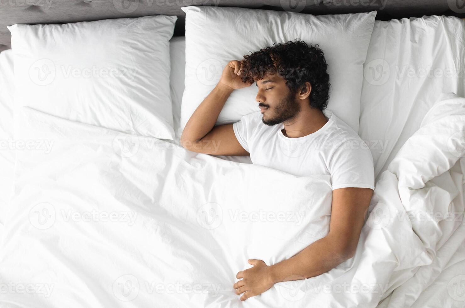 Top view of handsome bearded young hindu guy in white t-shirt resting at home, sleeping alone in his comfortable king size bed, panorama with copy space. Healthy sleeping concept photo