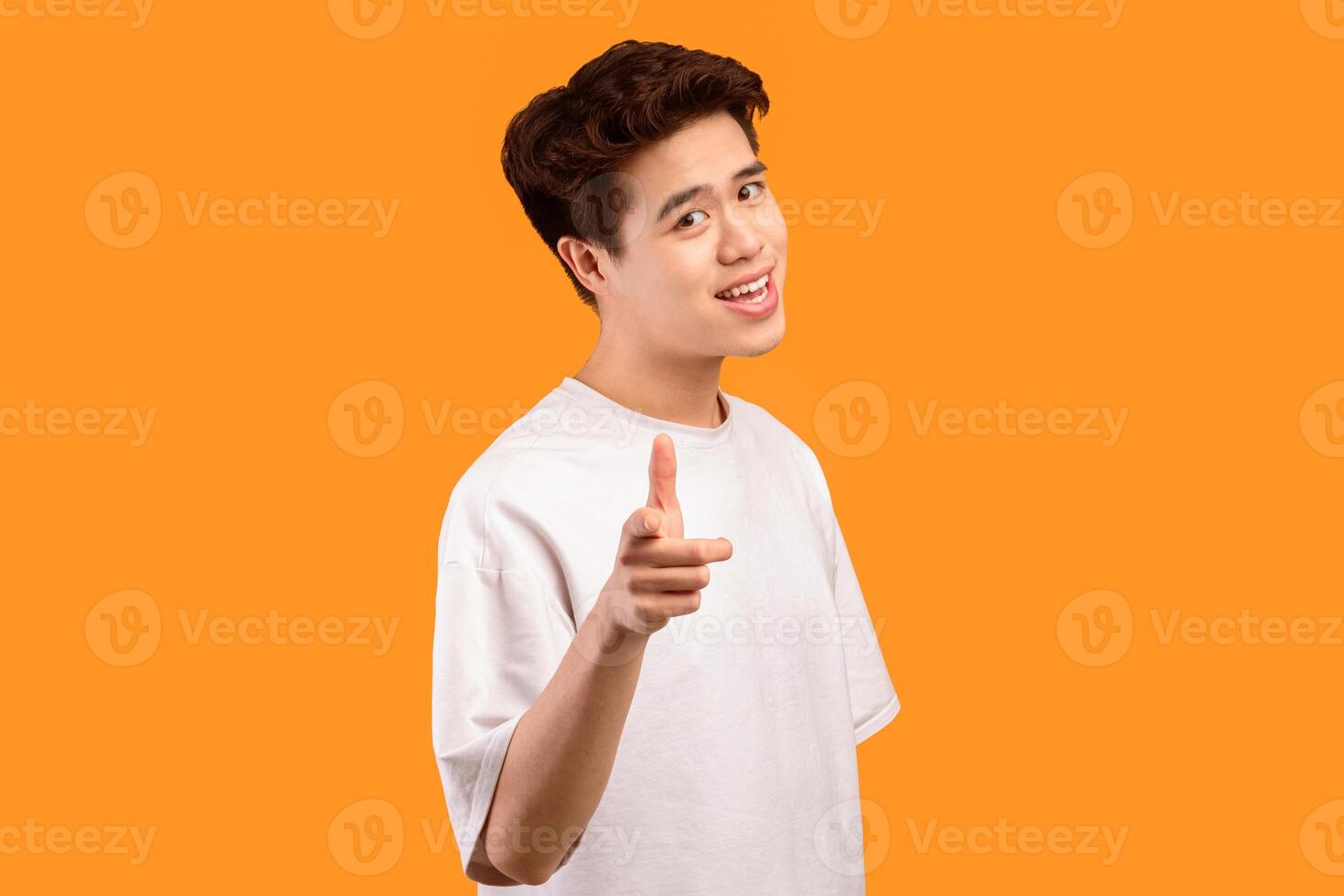 I Pick You. Portrait of casual asian guy in white t-shirt pointing finger at camera, posing isolated over orange studio background wall. Positive smiling man choosing and indicating photo
