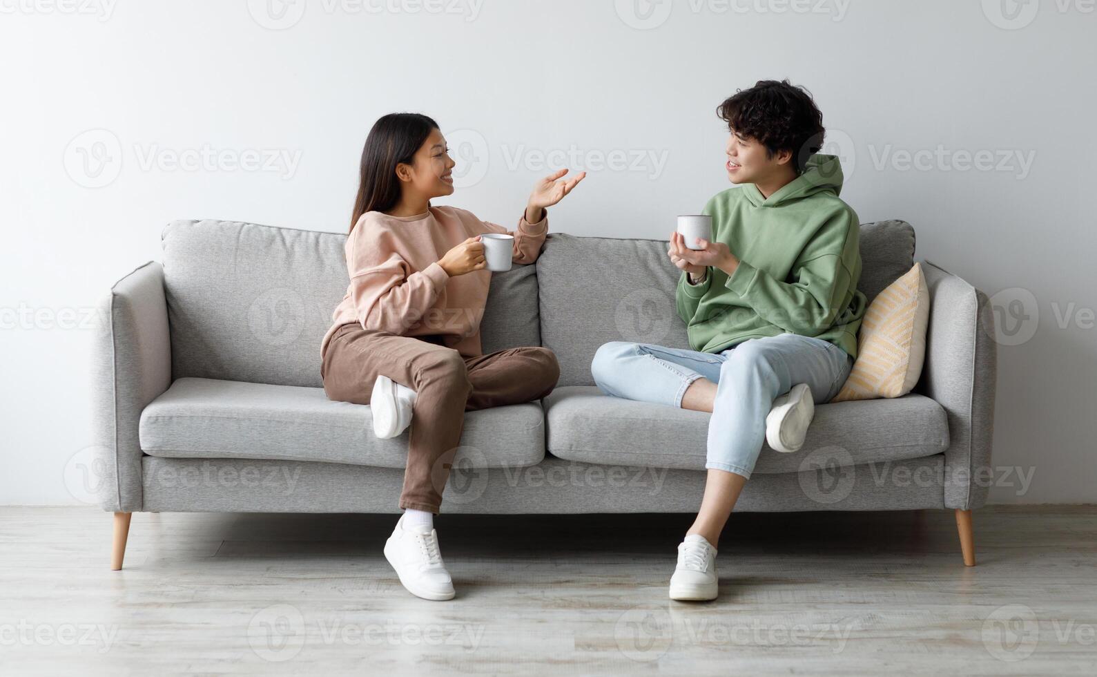 Young Asian boyfriend and girlfriend drinking coffee and chatting while sitting on sofa at home. Affectionate millennial couple enjoying hot drink, having conversation, spending time together photo