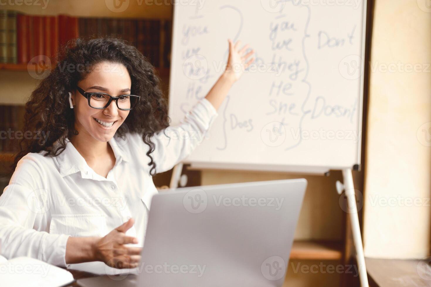 Online Education. Female English teacher having conference chat with students and class group, using laptop. Woman wearing glasses and wireless headphones, pointing at board with grammar rules photo