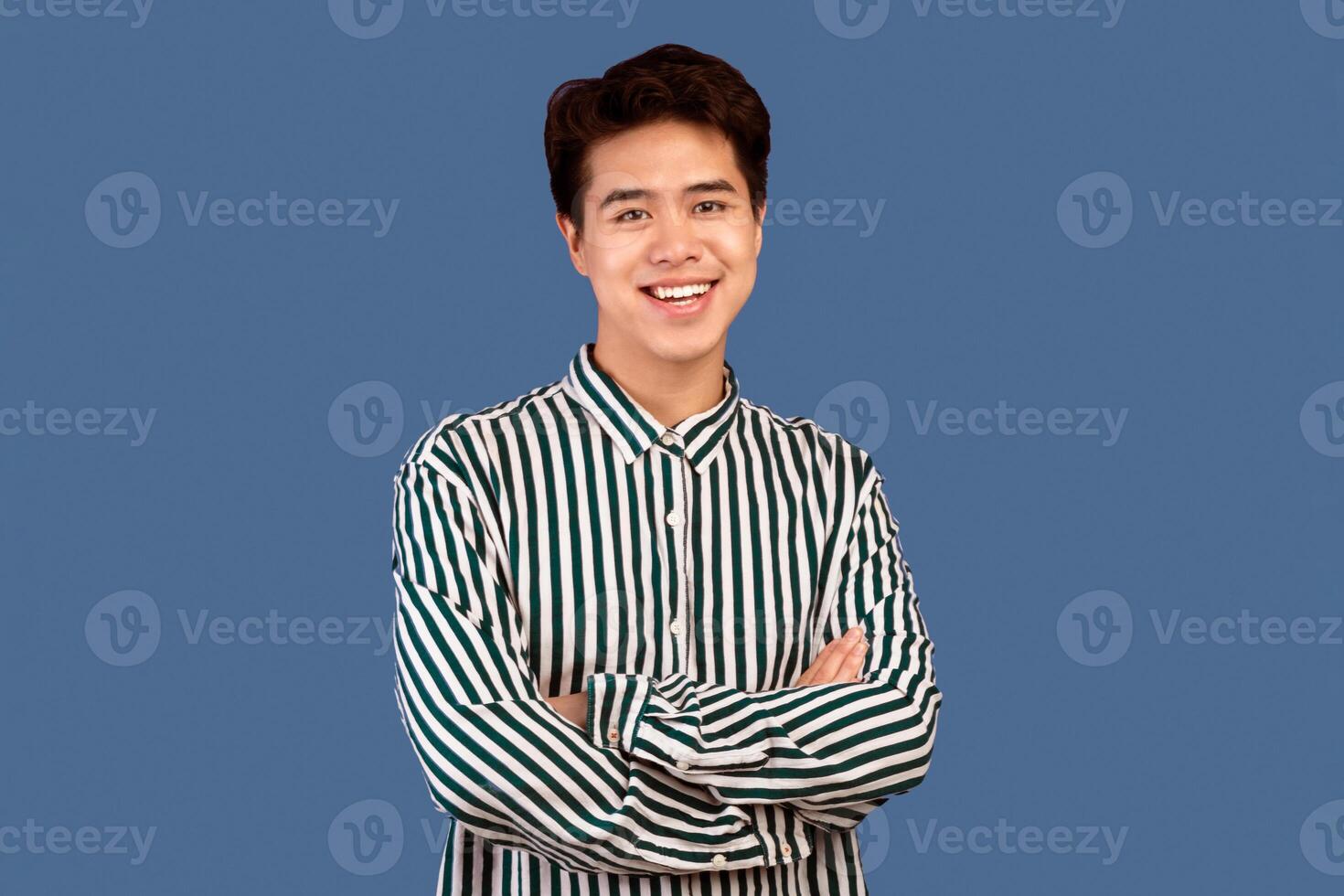 Casual Confident Person. Portrait of smiling asian guy with folded arms looking at camera, wearing striped shirt, standing isolated on purple studio background. Happy casual male teenage model posing photo