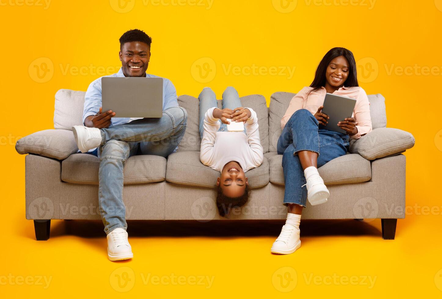 Gadgets Addiction. Young black family of three people holding and using different electronic devices sitting on sofa on yellow orange background. Parents and their daughter with modern technology photo