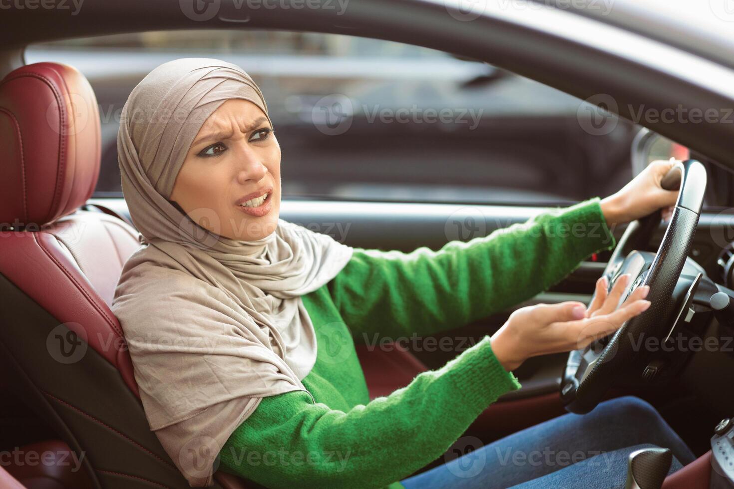 Angry muslim woman arguing with someone while driving car, sitting on front seat and emotionally gesturing. Annoyed arab lady stuck in traffic jam, having road accident, complaining about bad drivers photo
