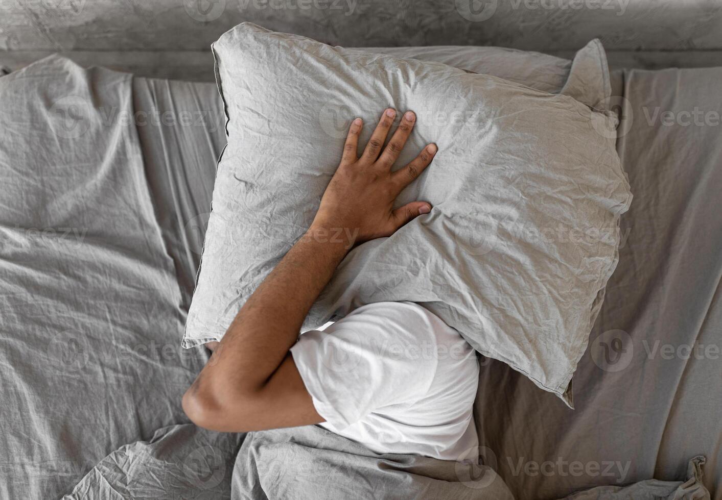 Closeup of irritated young black man lying in bed and covering head and ears with pillow, hearing and suffering from too loud sound. Angry millennial guy can't sleep, tired of noisy neighbors photo