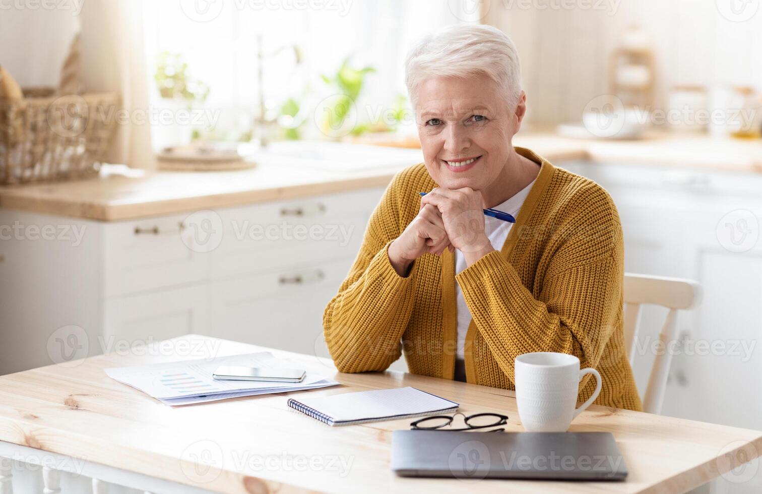 Positive elderly lady freelancer working from home or studying, sitting in kitchen with laptop and smartphone, taking notes while reading financial report, smiling at camera, copy space photo
