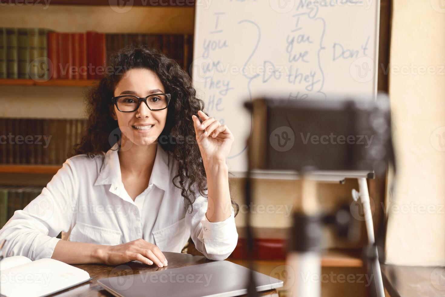 Online Lesson. Young female English language professor in glasses sitting at desk, teaching grammar rules, giving remote lecture. Woman recording blog for her internet channel using camera photo