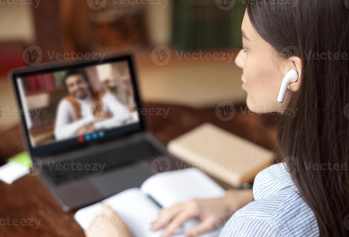 Online education, e-learning concept. Young woman with earbuds having conference with male teacher, sitting at desk and using laptop, taking notes, discussing studying material, home interior photo