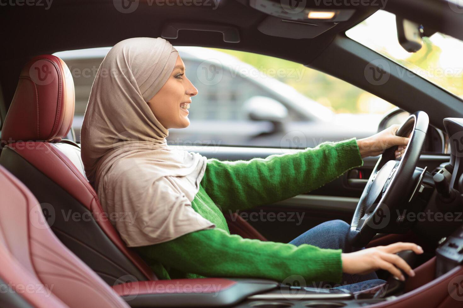 Female Driver. Side View Portrait Of Smiling Islamic Lady In Headscarf Learning To Drive Luxury Automobile In Urban City, Sitting On Driver's Seat, Holding Steering Wheel And Gear Shift photo