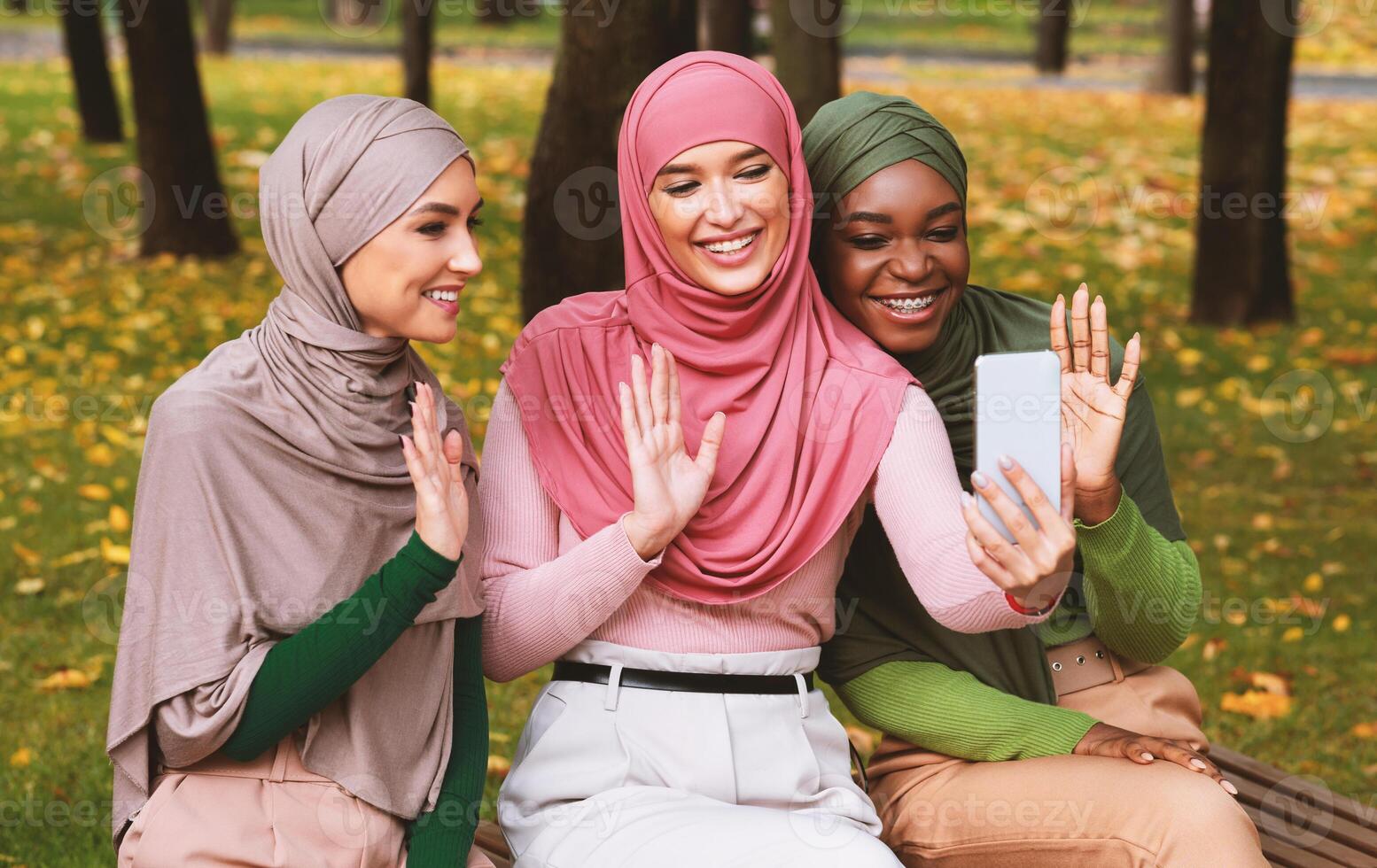 Three Muslim Ladies Calling Waving Hello To Smartphone On Bench In City Park Outdoors. Islamic Females Friends Chatting Making Call. E-Learning, Modern Remote Communication Concept photo