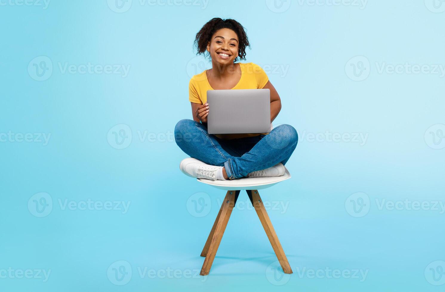 Cheery young black woman working online, sitting on chair and using laptop on blue studio background, full length. Cheerful African American lady surfing internet on portable pc photo