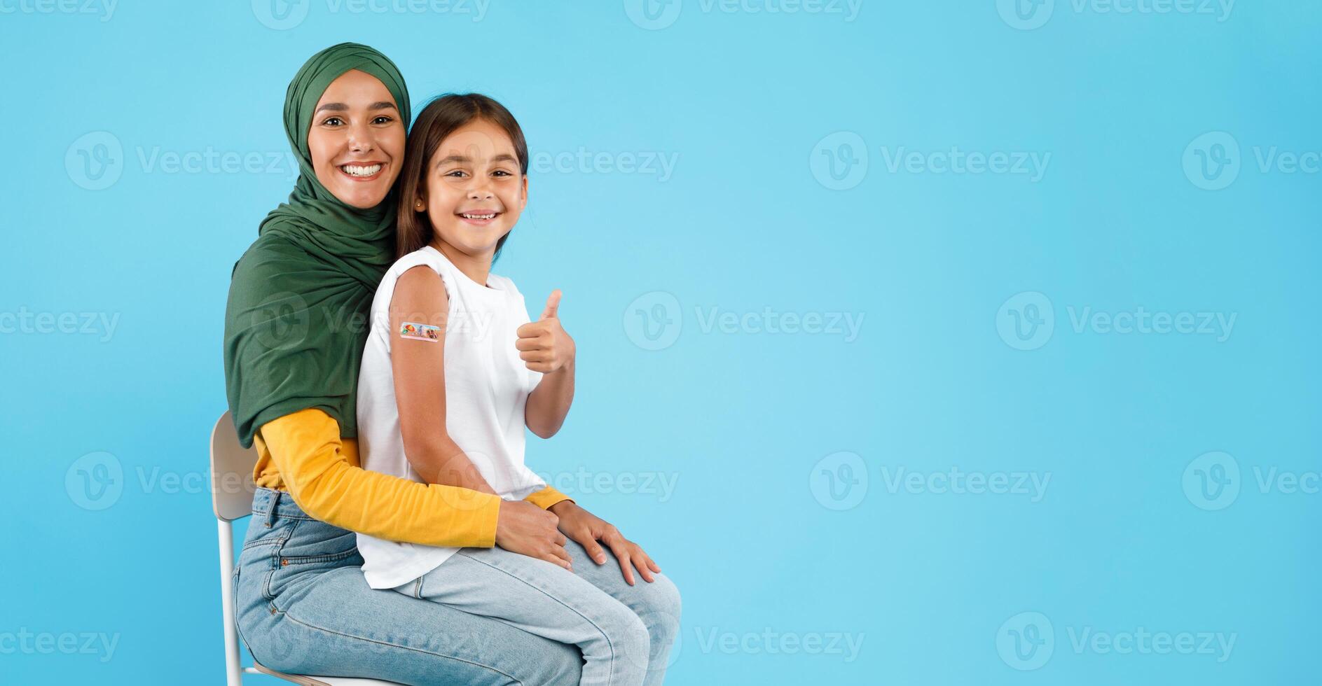 Covid-19 Vaccination For Children. Vaccinated Muslim Mother And Little Daughter With Plaster On Arm After Vaccine Injection Gesturing Thumbs Up Sitting On Blue Background. Panorama, Copy Space photo