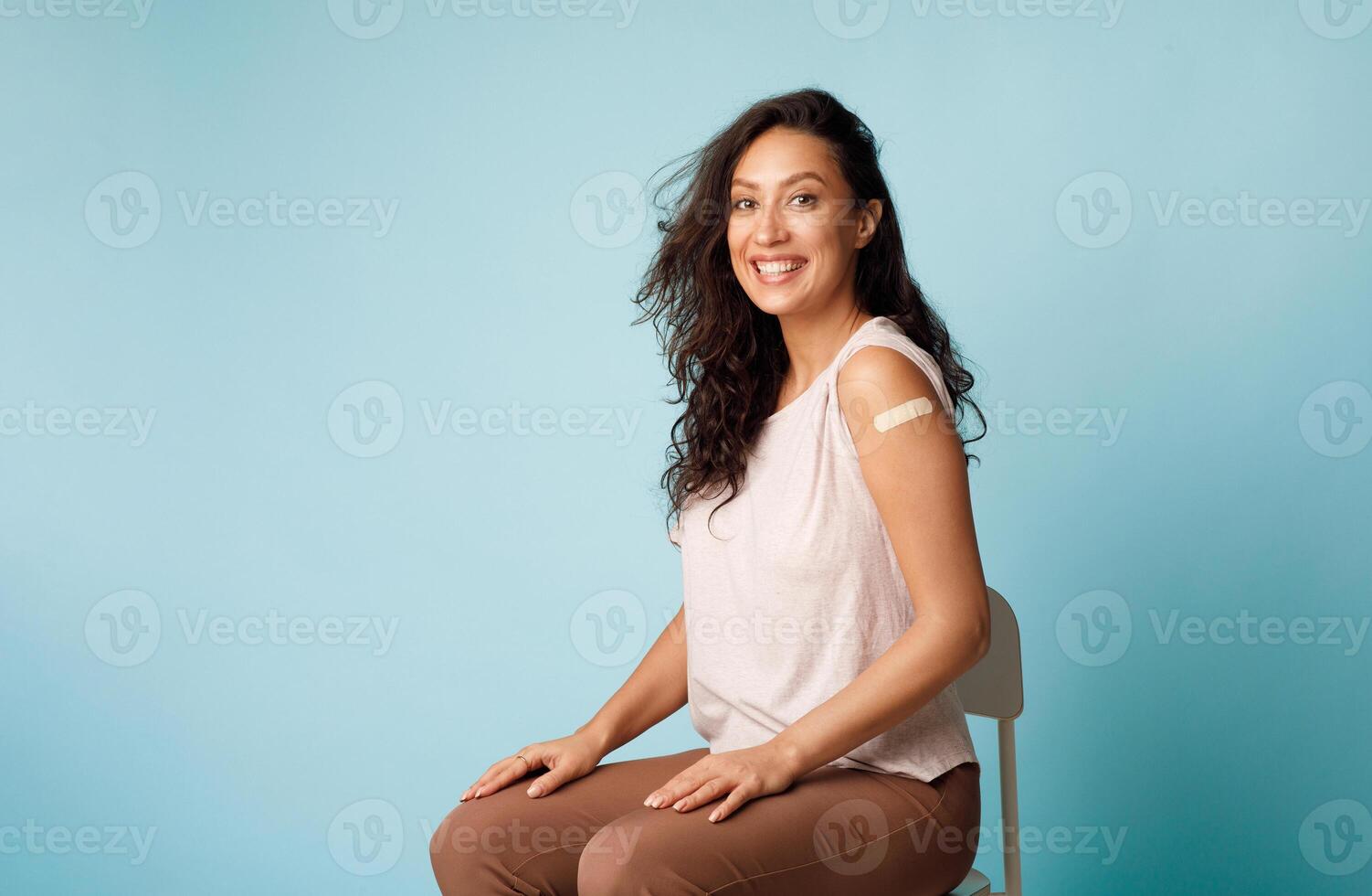Covid-19 Vaccination. Happy Young Woman Showing Vaccinated Arm After Getting Coronavirus Vaccine Injection With Plaster Strip Sitting Posing On Blue Background, Smiling To Camera. Covid Immunization photo