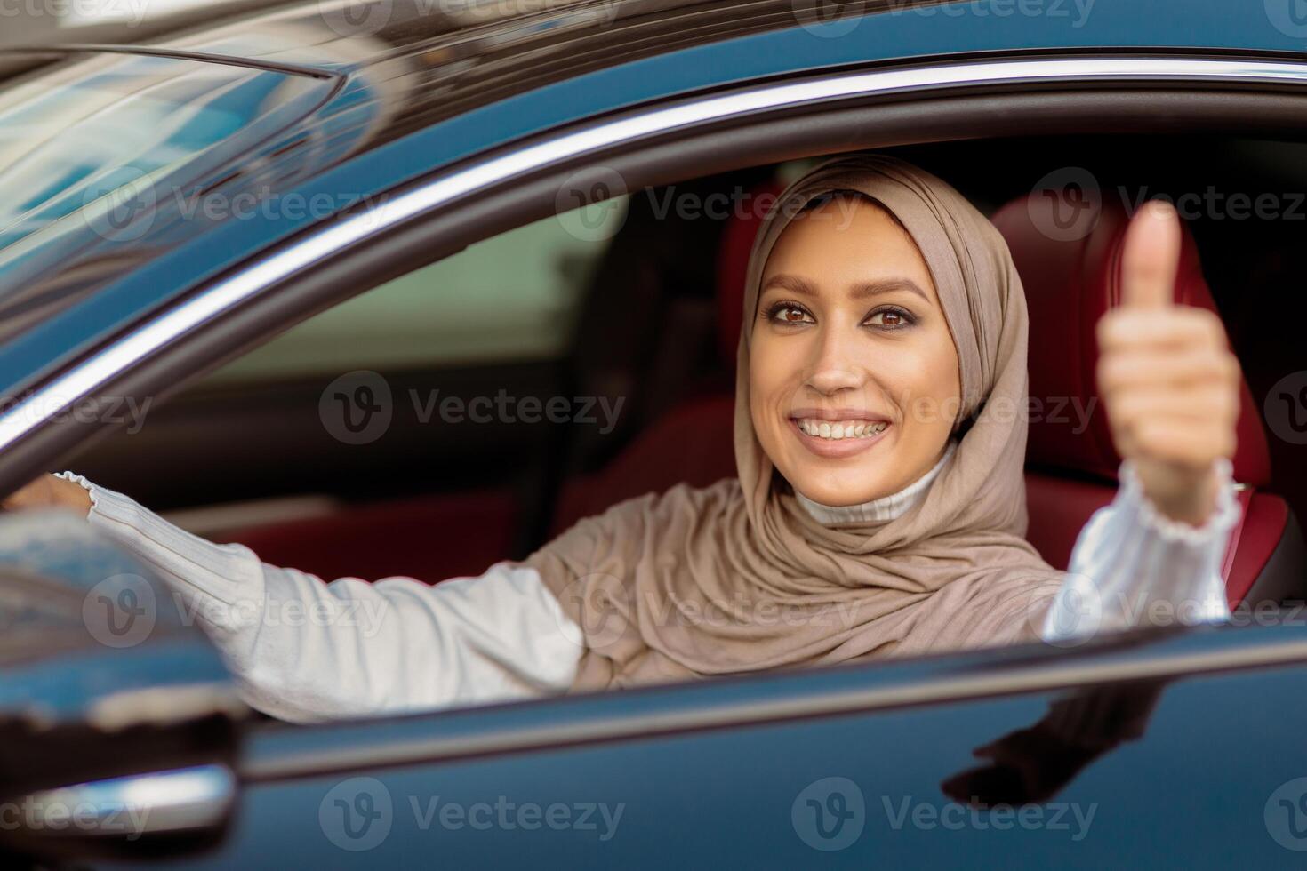 Happy Car Owner. Portrait Of Positive Middle East Lady In Hijab Showing Thumbs Up, Sitting In Driver's Seat, Happy About Purchase In Dealership Showroom. Excited Muslim Woman Making Approval Gesture photo
