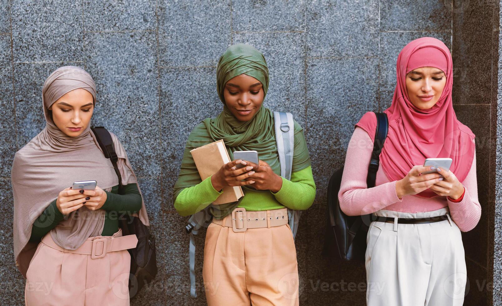 Three Modern Muslim Women Using Cellphones Texting And Networking Standing Over Gray University Building Wall Outdoor. Islamic Students Ladies With Smartphones Using Mobile Applications photo