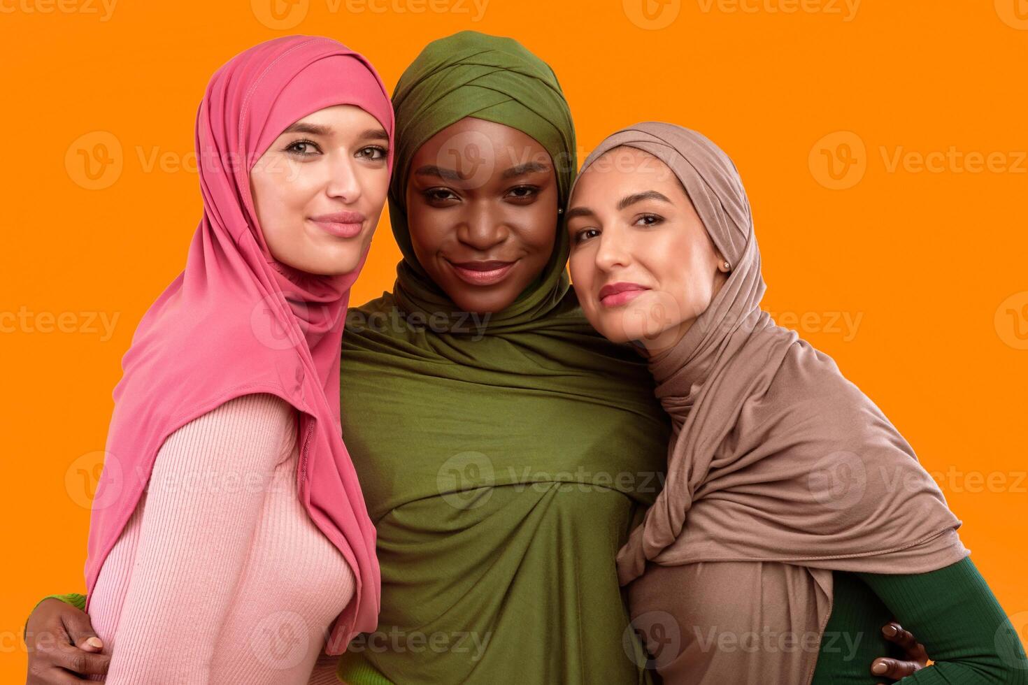 Three Muslim Women In Hijab Headscarf Embracing Posing Smiling To Camera Standing Together In Studio On Yellow Background. Modern Islamic Ladies Portrait. Arabic Female Fashion Concept photo