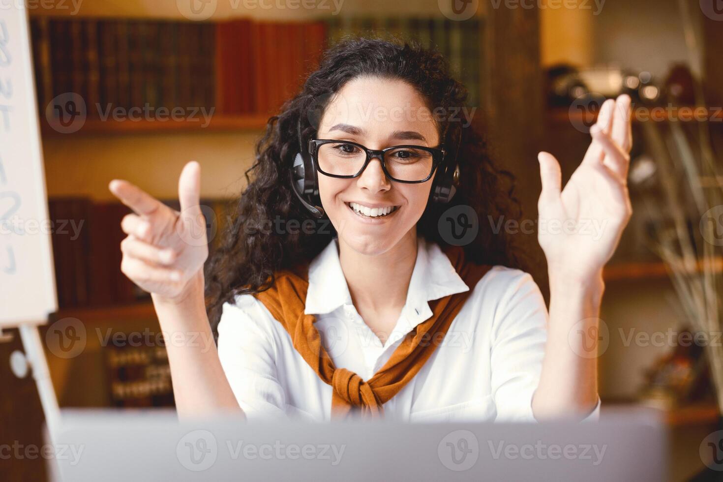 Virtual Tutoring. Positive Young Female Professor Wearing Eyeglasses And Wireless Headphones Having Call, Teaching New Theme Or Subject, Talking To Camera During Online Lesson At Home photo