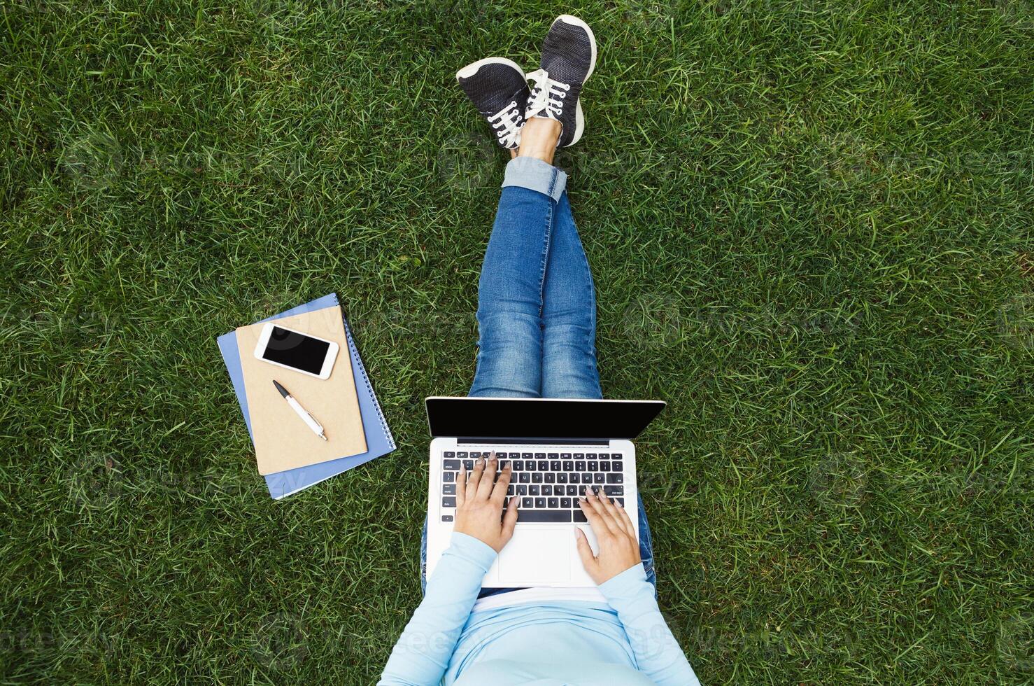 Top view of young woman in casual outfit using laptop while sitting on grass with notebook and smartphone. Concept of freelance job or study, copy space photo