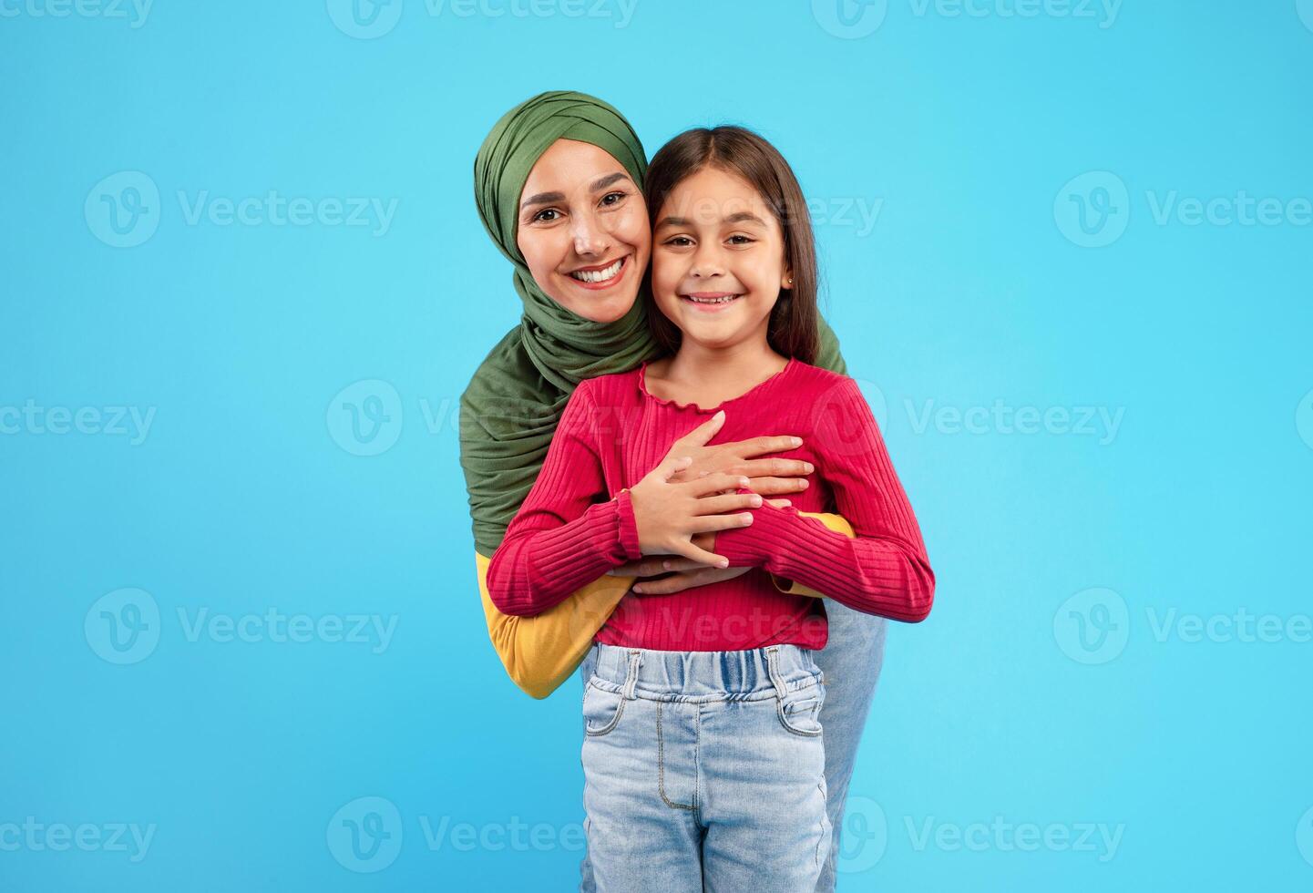 Cheerful Islamic Mother Wearing Hijab Hugging Her Little Daughter Smiling To Camera Posing On Blue Background. Arabic Mom And Her Kid Girl Standing And Embracing Together In Studio photo
