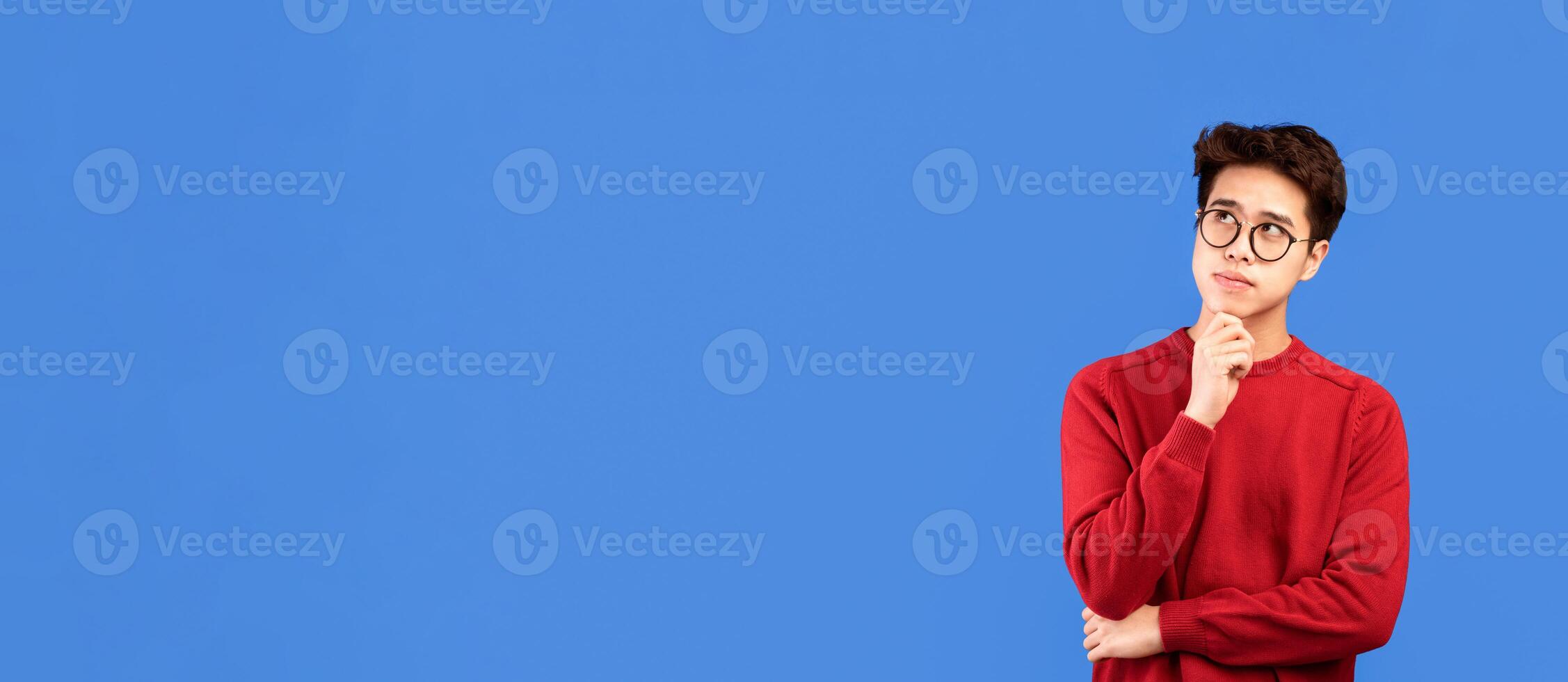 Good Offer. Portrait of pensive asian man in glasses looking up at copy space, touching his chin, isolated over blue studio background. Thoughtful teen guy thinking and making decision photo