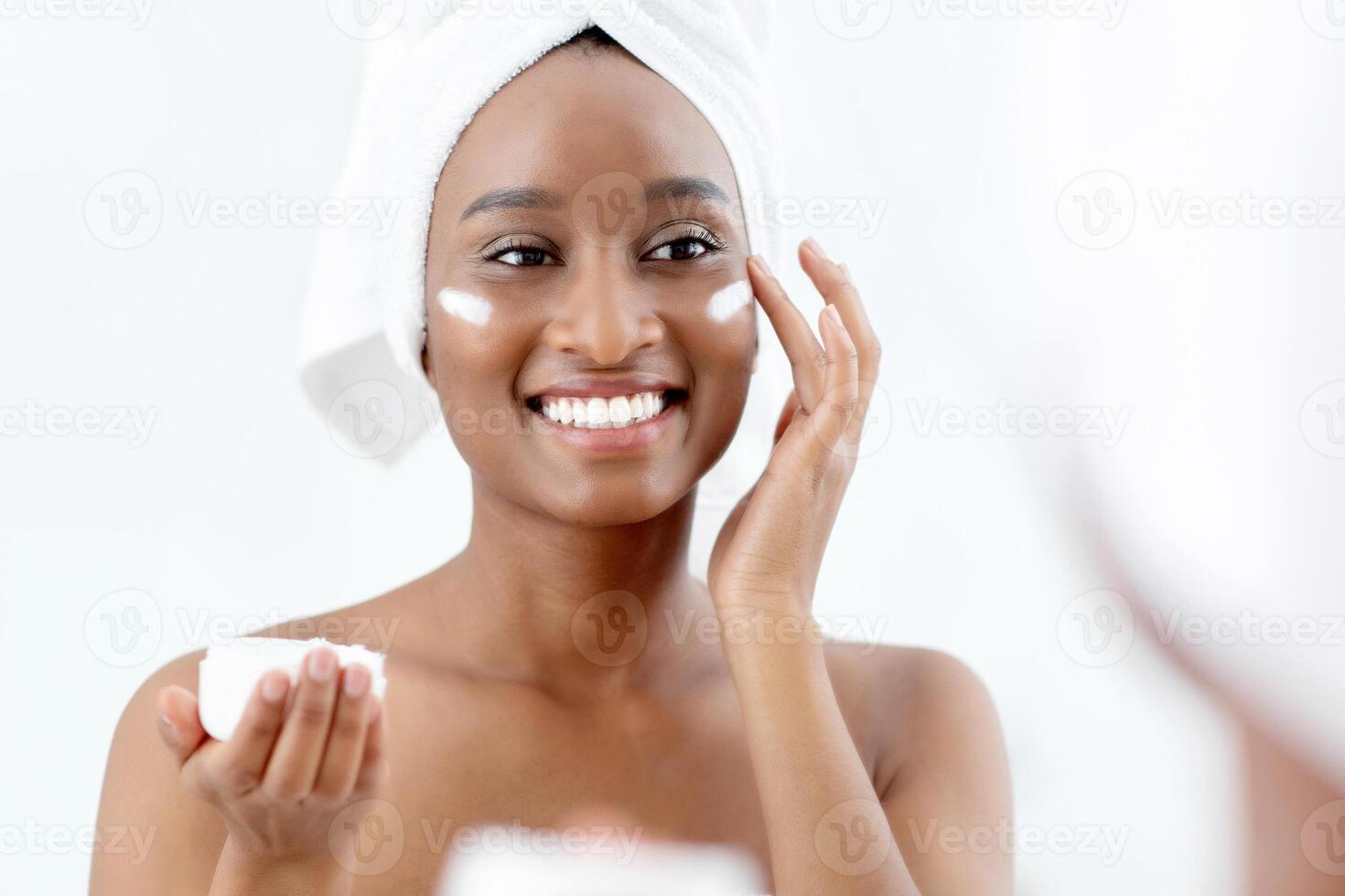 Anti-wrinkles, chemical peeling, anti-ageing treatment. Smiling cute millennial african american lady with towel on head after shower applying cream on face and hold jar in hand, in bedroom in morning photo