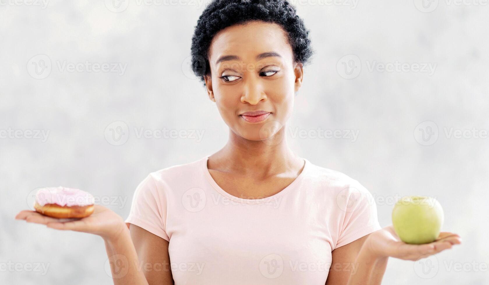 Healthy diet vs junk food. Pretty black lady holding apple and donut in her hands, tempted to eat dessert instead of fruit. Young woman having difficulty choosing good meal over unhealthy one photo