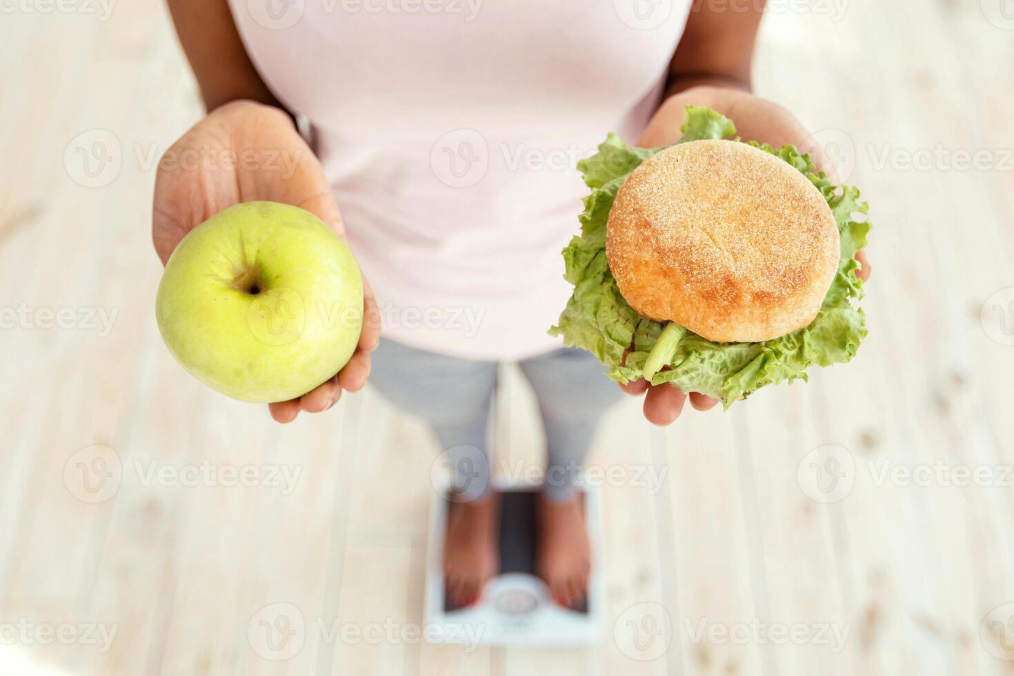 Above view of black woman holding apple and hamburger, standing on scales, making choice of her diet, closeup. African American lady choosing healthier nutrition for wellbeing and weight loss photo