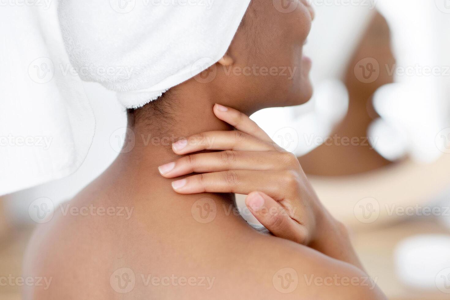 Beauty and spa day at home at weekends and free time. Pretty fresh millennial african american woman in white towel smears her body with cream after shower, in interior of bedroom in morning, back photo
