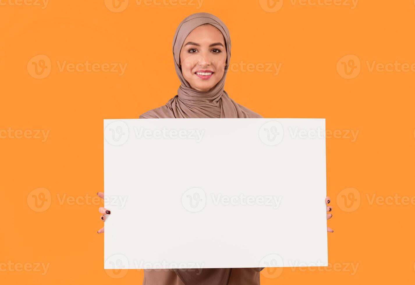 Muslim Lady Holding Blank Poster Board For Text Advertisement Posing Over Yellow Studio Background, Wearing Hijab Headscarf. Islamic Woman Advertising Something With Empty Paper Sheet Banner. Mockup photo