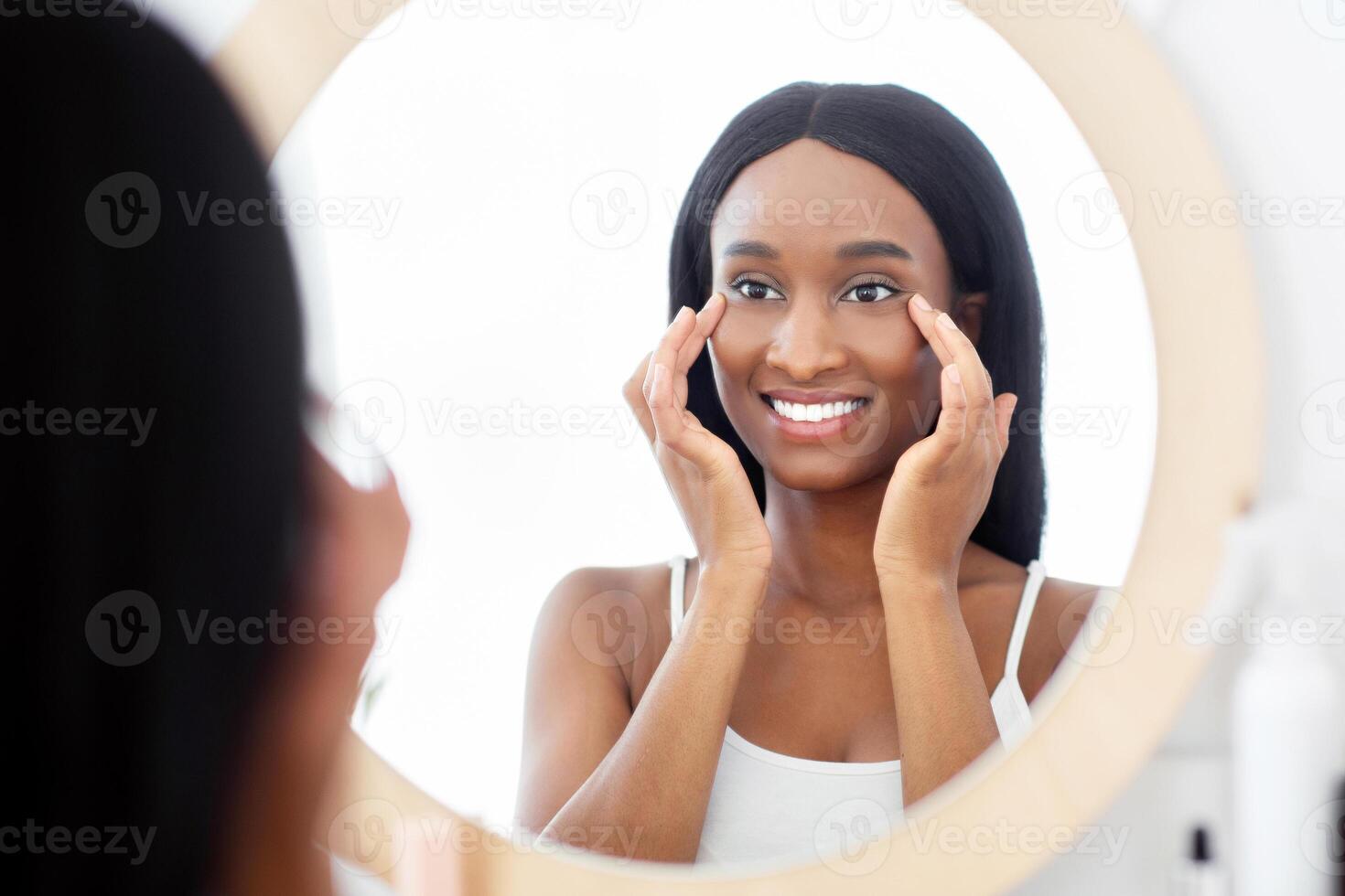 Beauty needs care and home spa during covid-19 quarantine. Smiling young african american woman smears face with cream, look at mirror and fights wrinkles and looks in mirror in bedroom interior photo