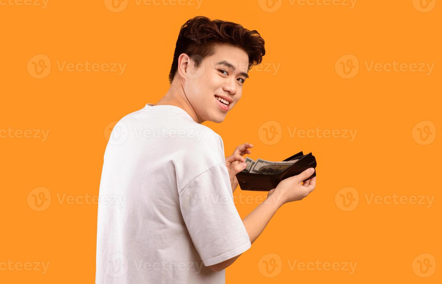Finance, Investment And Money Saving. Portrait of excited asian young man holding and showing wallet full of dollars, free space, isolated over orange studio background. Greedy guy got salary photo