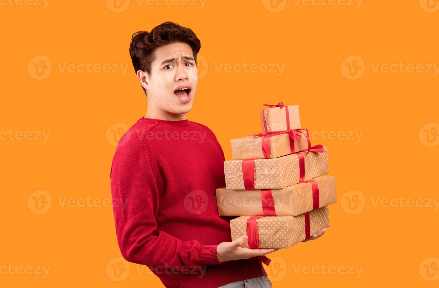 Time For Celebration. Portrait of emotional asian guy holding stack of present boxes isolated over orange background. Excited man standing with pile of gifts wrapped in craft paper and red ribbons photo