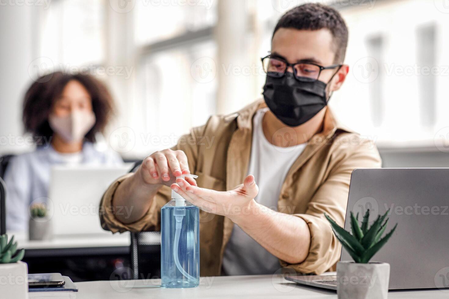 Disinfection and social distancing in office during COVID-19 epidemic. Young man worker in protective mask cleans his hands with antiseptic at workplace and gets to work with laptop, free space photo