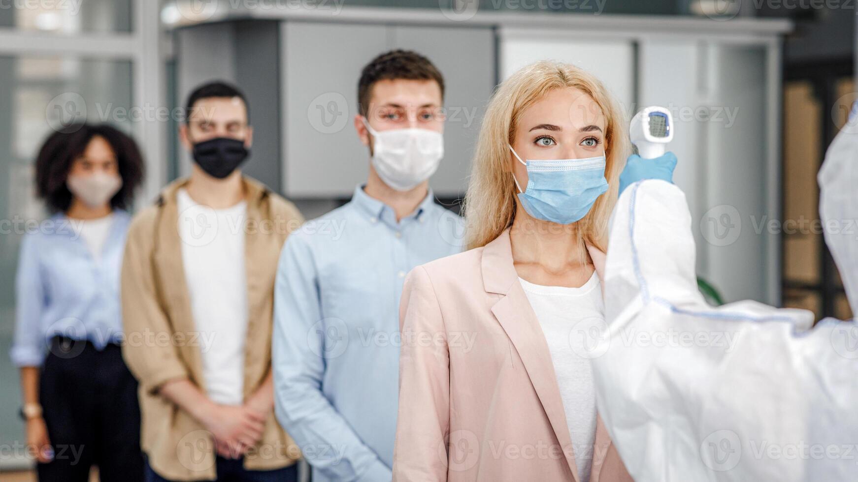 Temperature control test at work and social distancing. Man in protective suit checks multinational workers with non-contact thermometer in morning at entering in office, empty space photo