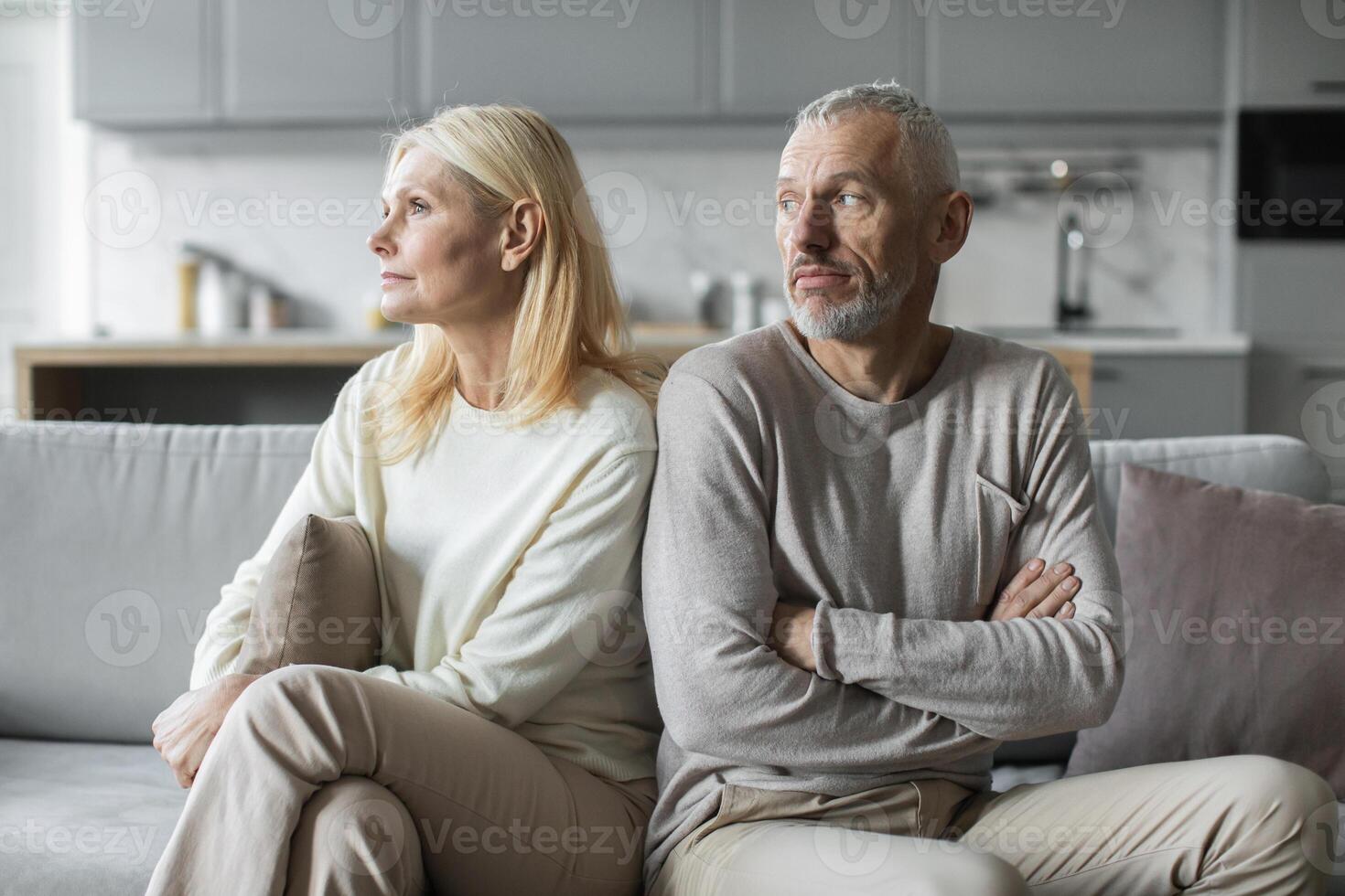 A senior man and woman sit at opposite ends of a couch in a modern living room, both appearing lost in thought and reflecting on their unresolved issues, highlighting emotional distance. photo