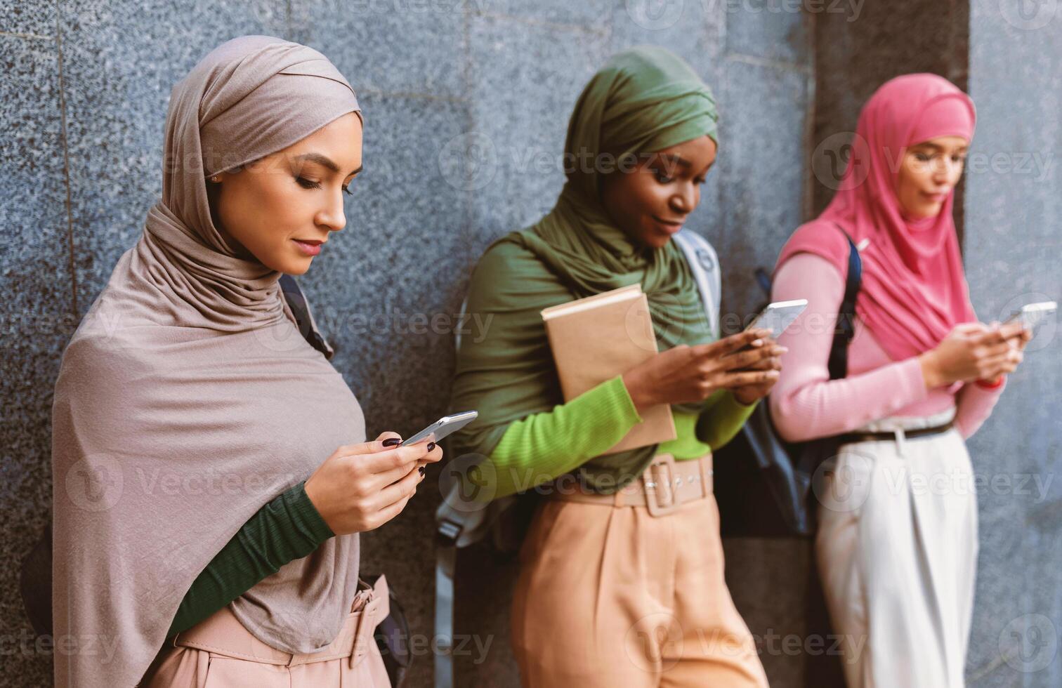 Modern Arab Ladies Using Smartphones Mobile Apps Texting And Networking In Social Media Standing Over Gray Wall Outside. Female Muslim Students With Cellphones Using Applications photo