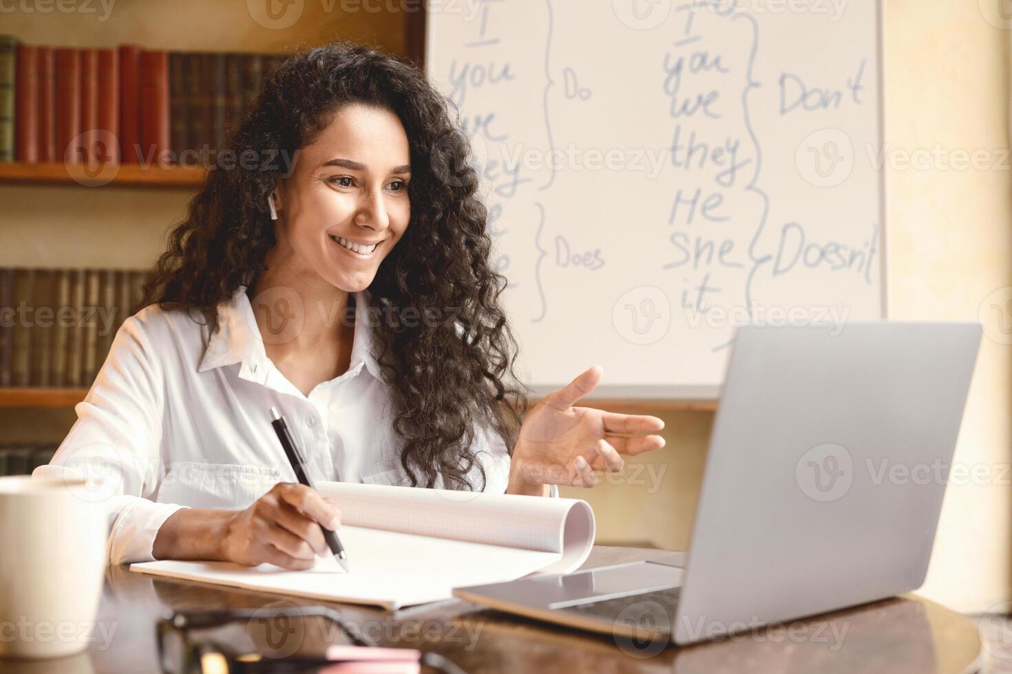 E-learning And Online Course During Quarantine. Positive young female teacher in earphones having online call with students using laptop, writing and taking notes in her notebook, talking to webcam photo
