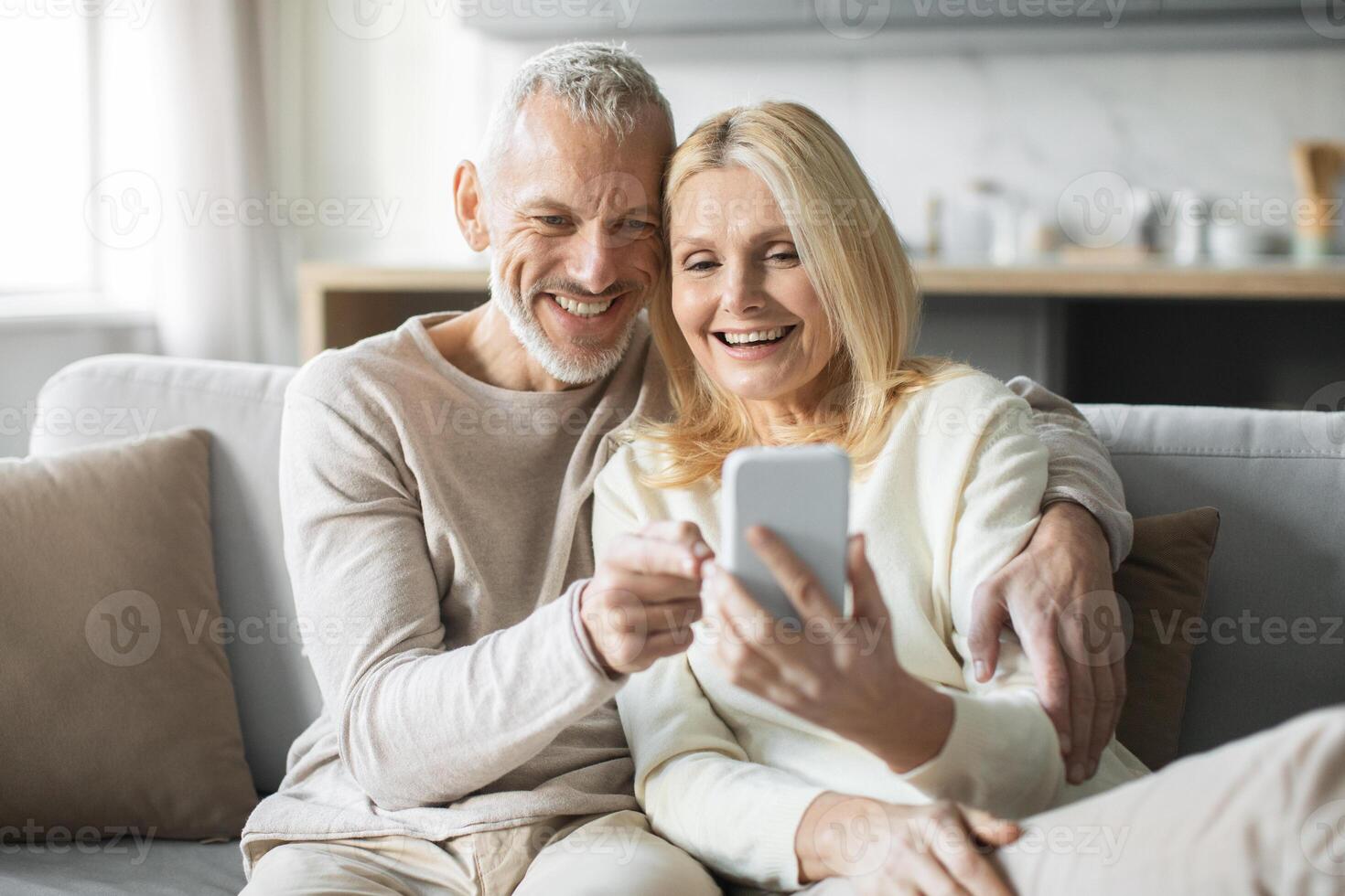 A happy senior couple sits on a couch, smiling as they share a moment while looking at a smartphone. Their expressions reflect joy and connection, showcasing their bond in a cozy living room. photo