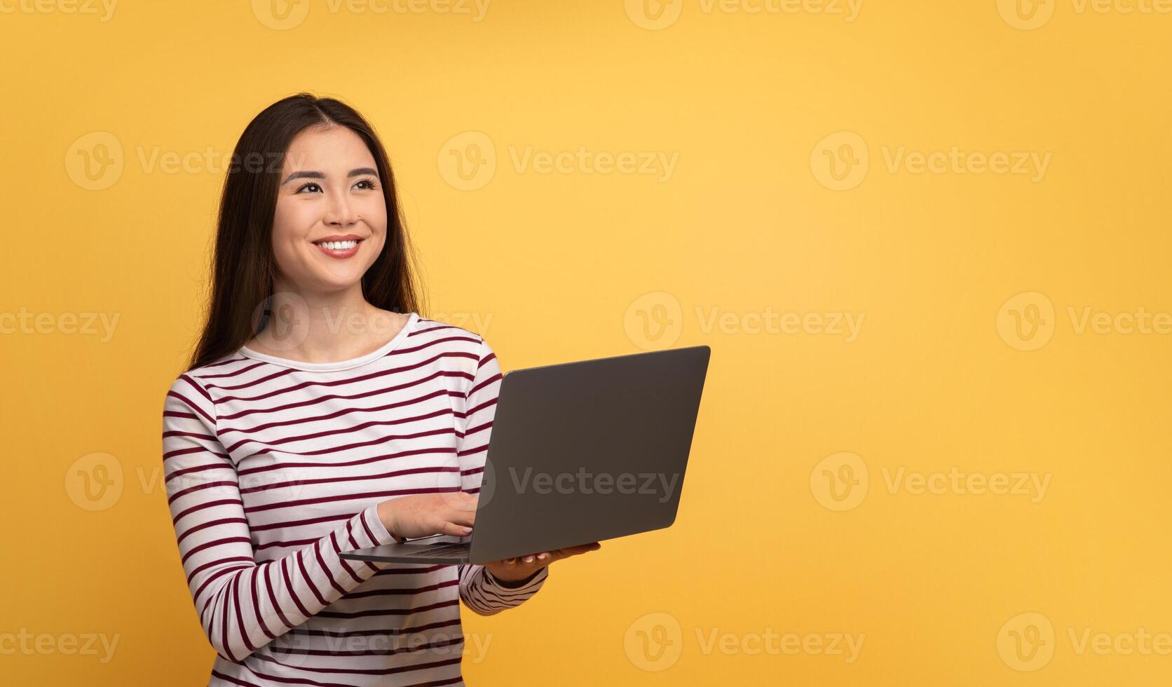 A woman with long hair holds a laptop and smiles cheerfully, showcasing a pleasant moment of engagement with technology. The bright yellow backdrop adds a lively atmosphere, copy space photo
