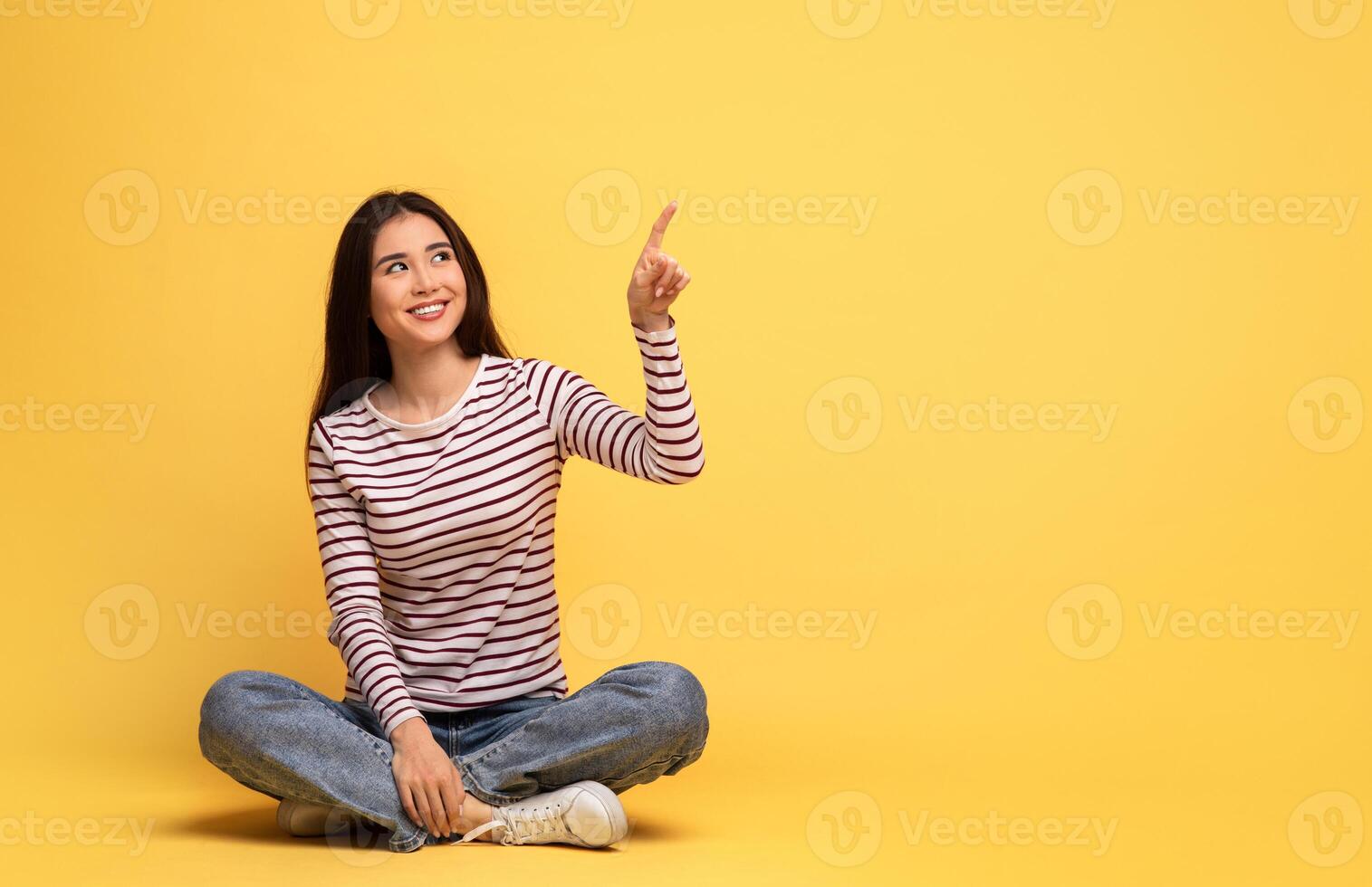 Asian young woman with long hair sits cross-legged on a bright yellow background, smiling and pointing upwards at copy space with one hand, suggesting excitement or inspiration. photo