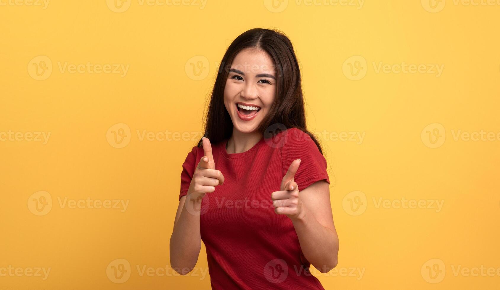 A cheerful young Asian woman with long hair smiles widely while playfully pointing her fingers at the camera. She stands against a vibrant yellow backdrop, exuding a fun and lively energy. photo