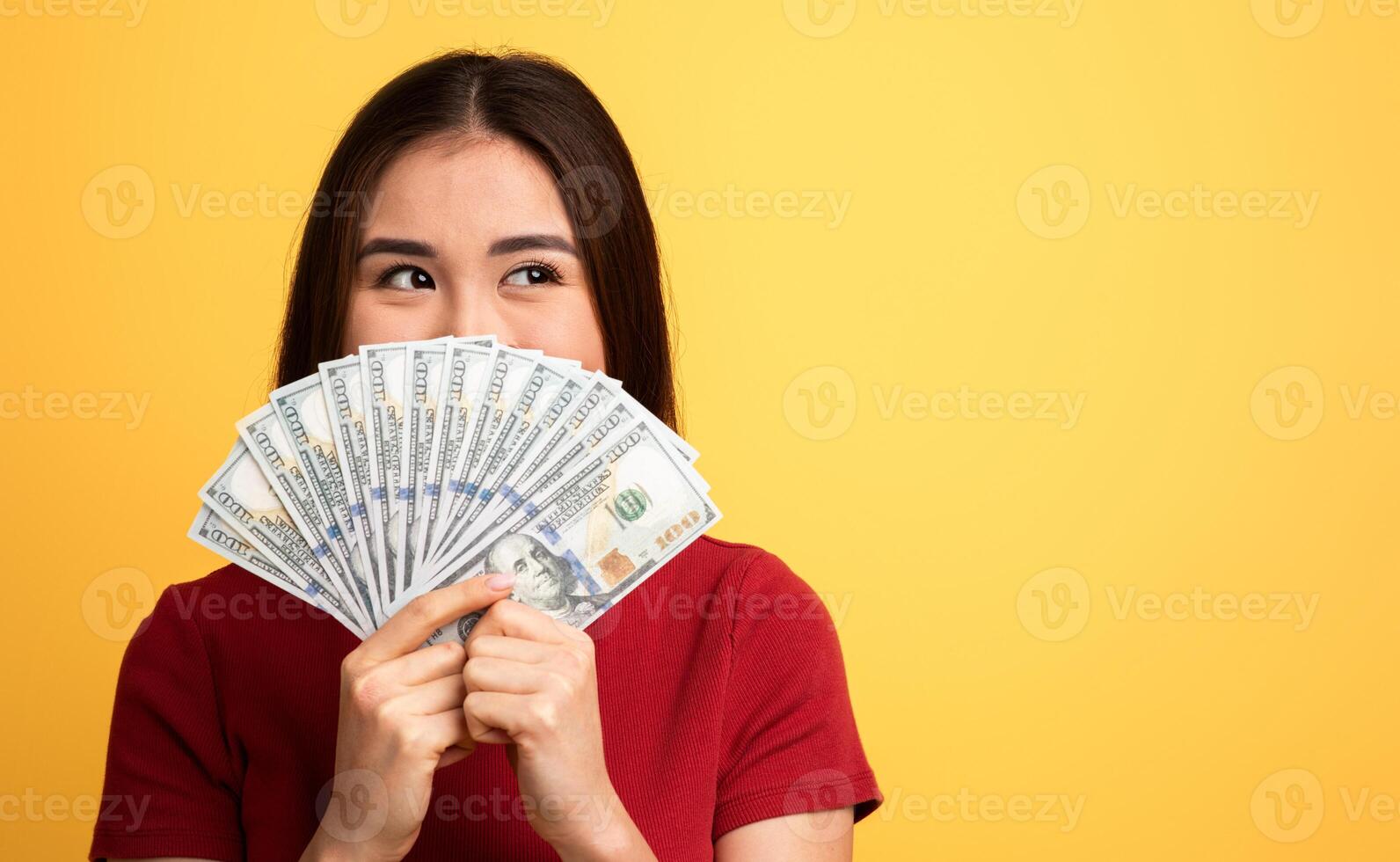 A young woman smiles while playfully holding a fan of hundred-dollar bills against a vivid yellow background. She seems excited and cheerful about her money, copy space photo