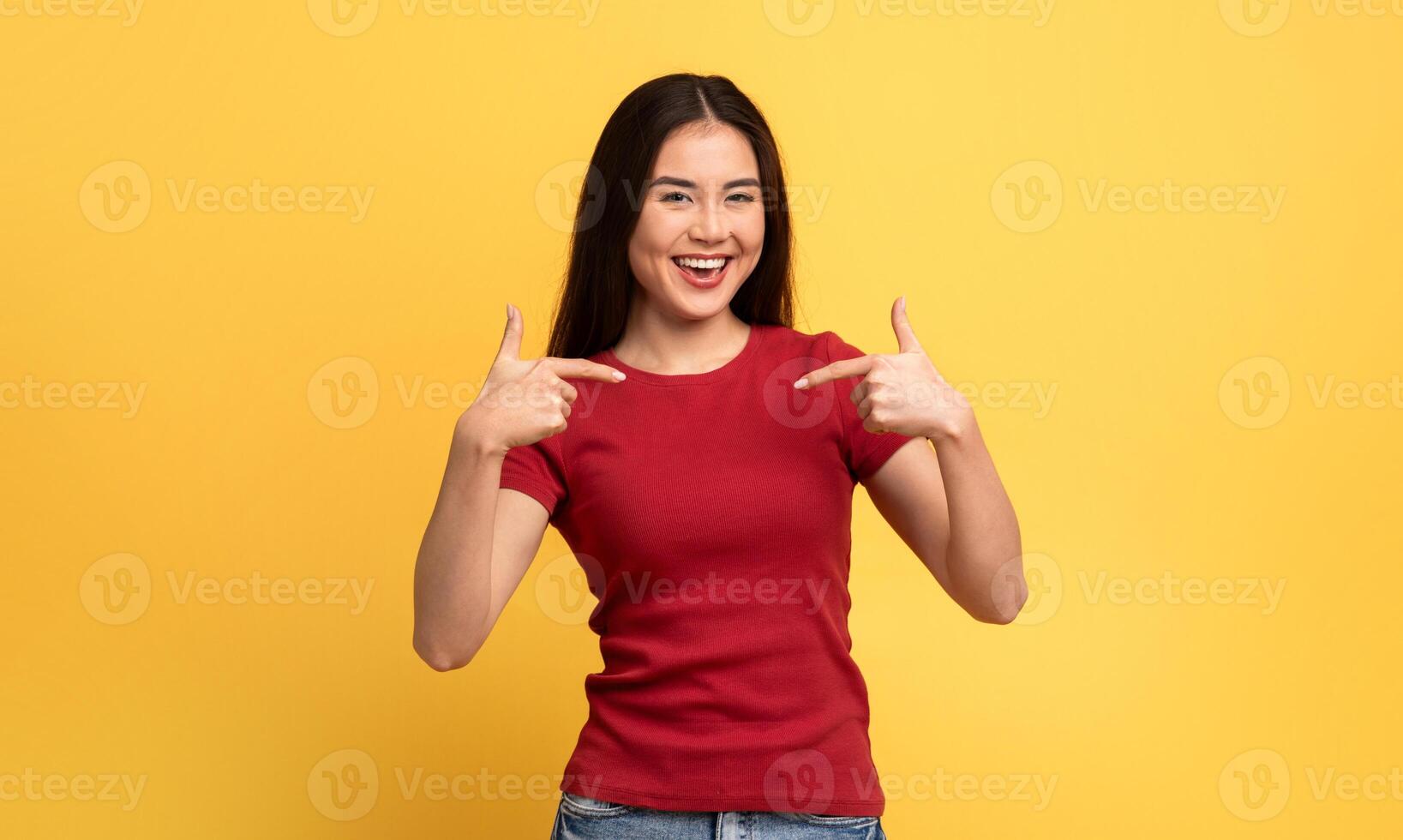 The setting features a young Asian woman joyfully pointing at herself, conveying a sense of excitement. She wears a red shirt that contrasts with the vibrant yellow backdrop. photo