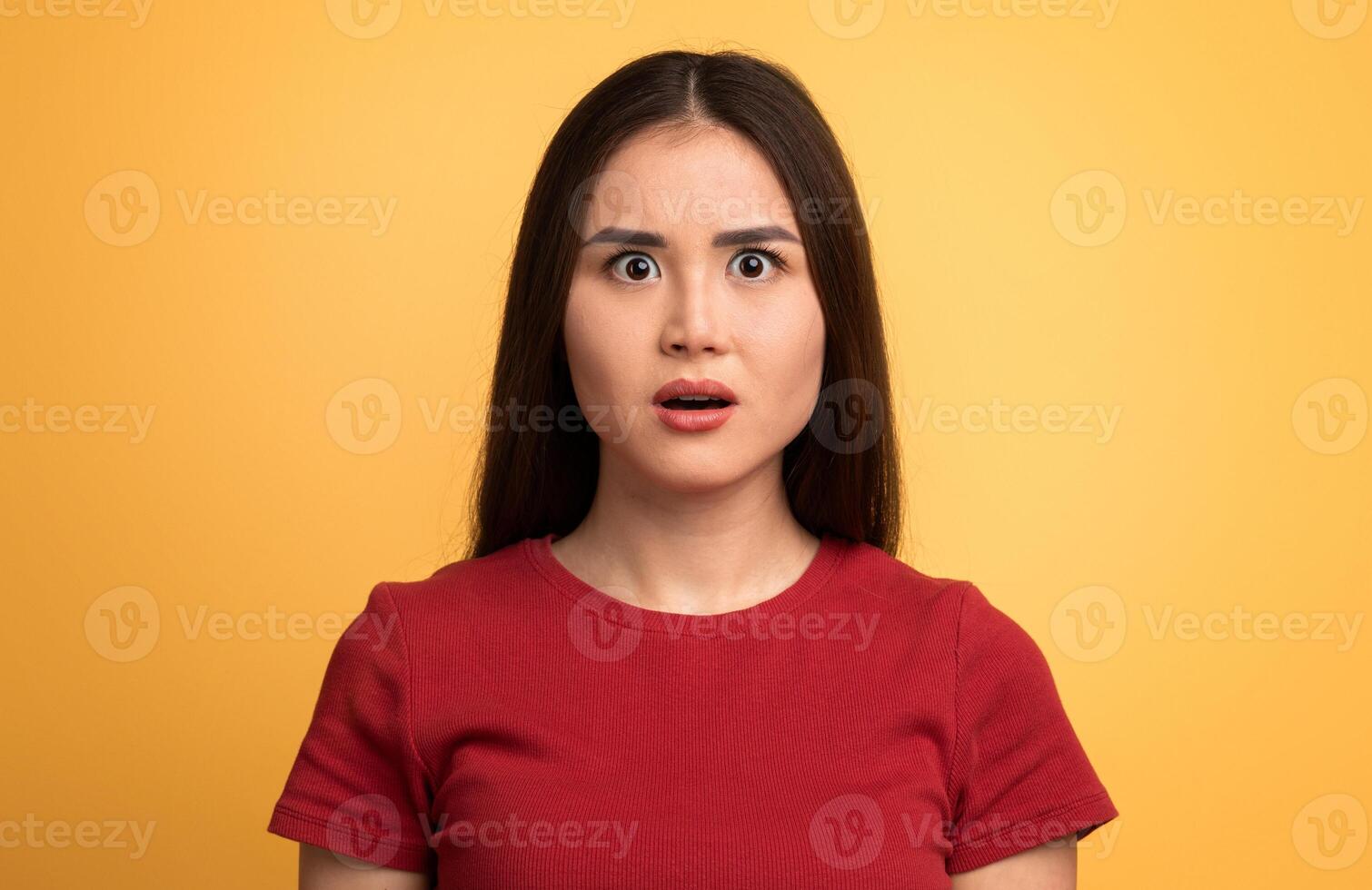 A young woman stands with her mouth slightly open, eyes wide in disbelief. She has straight long hair and wears a red shirt, set against a bright yellow backdrop. photo