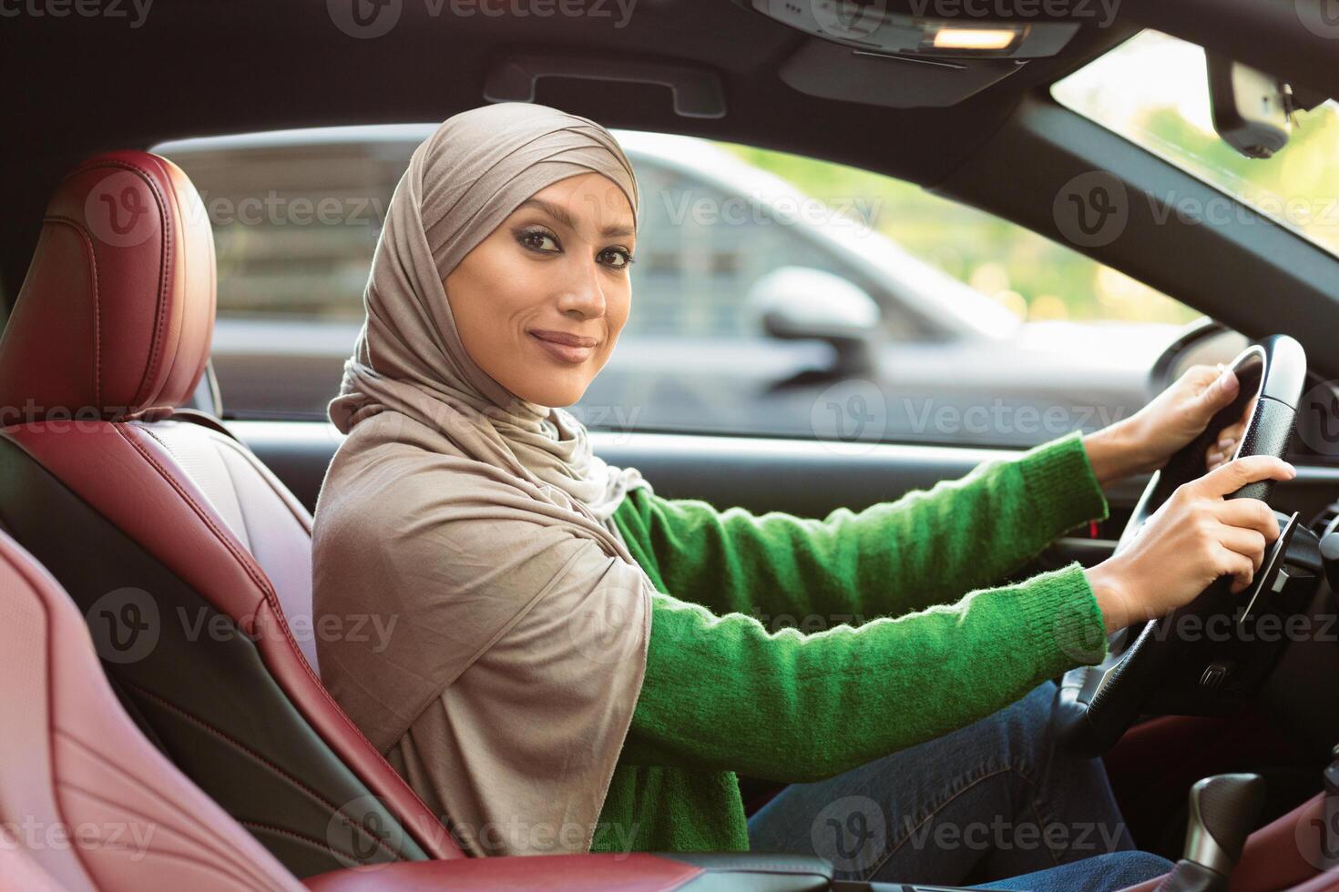 Vehicle Insurance Concept. Young muslim lady in hijab driving car, holding steering wheel, posing and looking in camera, smiling islamic woman in headscarf riding modern automobile in urban city photo