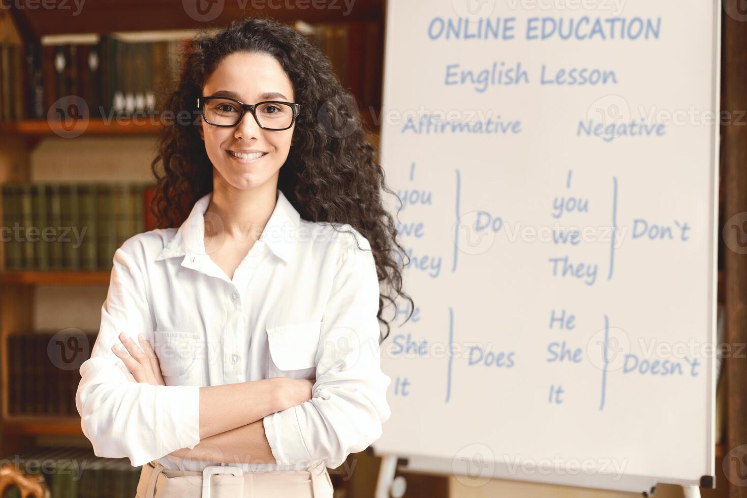 Online Education And E-Learning Concept. Portrait of smiling female teacher standing at whiteboard with English grammar rules. Confident woman in glasses looking at camera, posing with folded arms photo