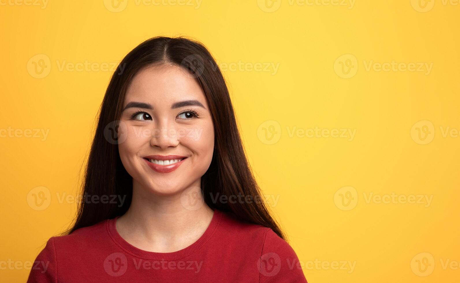 A young woman with long dark hair smiles playfully, looking to her side at copy space. The bright yellow background enhances her cheerful expression and adds warmth to the atmosphere. photo