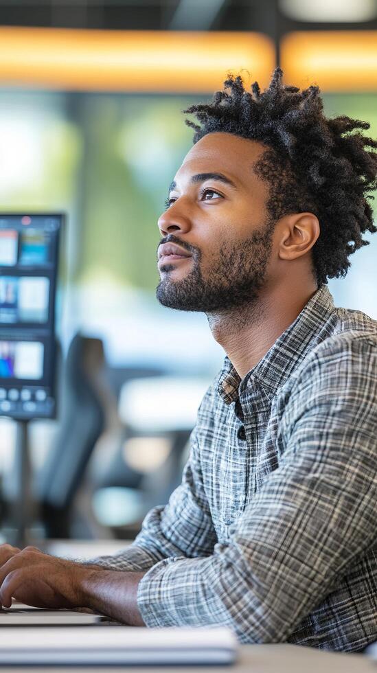 A thoughtful young African American man in a contemplative pose, working at his desk in a modern office setting. The image conveys focus, professionalism, and the atmosphere of a busy workspace. photo