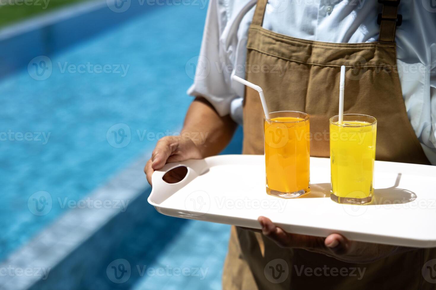 Waiter is carrying two glasses of ice tropical fruit juice to customer by the swimming pool in the luxury resort during summer for vacation and travel photo