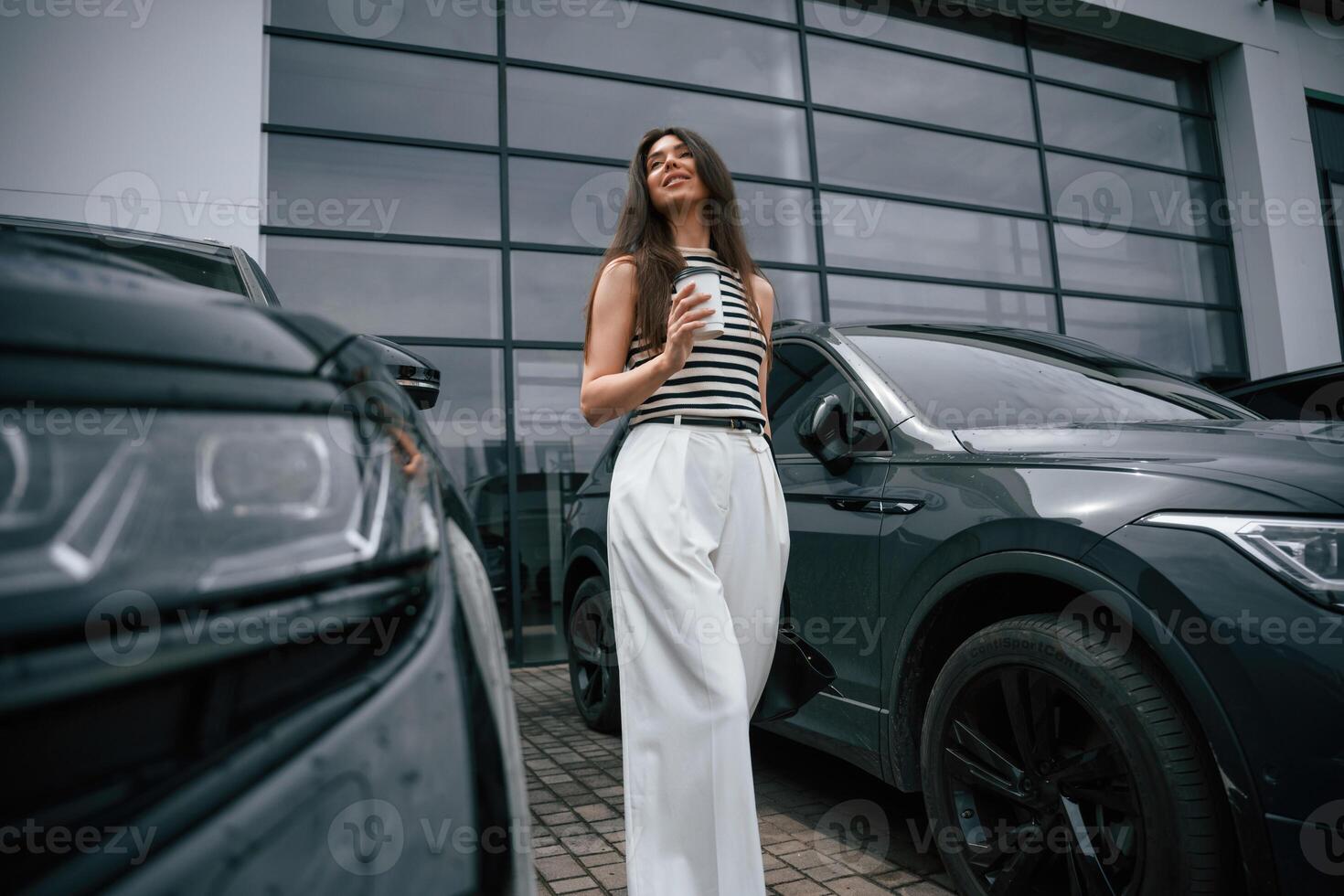 Holding cup of coffee. Attractive young woman is outdoors near her brand new car photo
