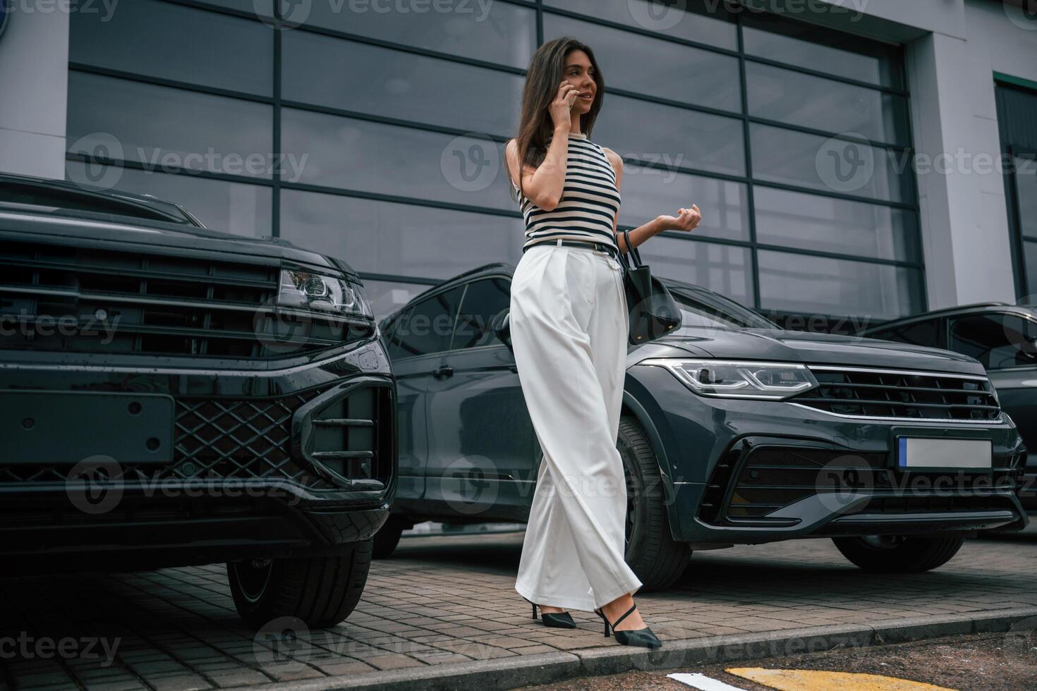 With, smartphone. Attractive young woman is outdoors near her brand new car photo